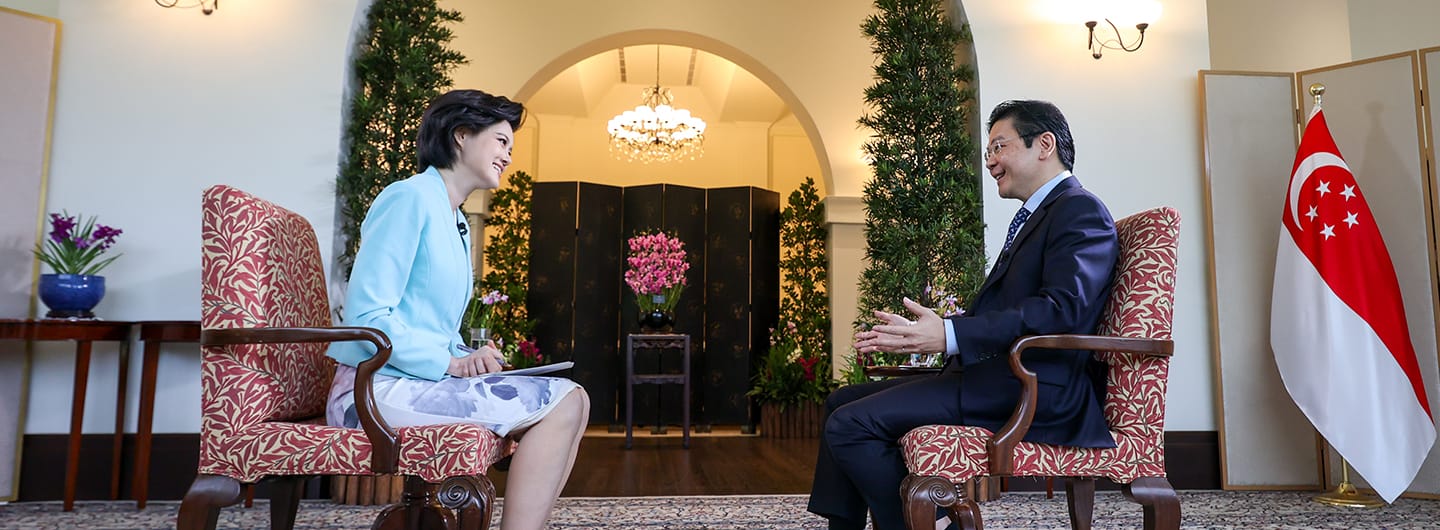 Interview setting: Woman in blue blazer, man in suit facing each other, Singapore flag.
